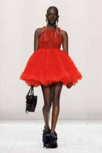 Model on a runway wearing a bright red textured mini dress with a voluminous, ruffled skirt, large silver hoop earrings, a black handbag, and black floral-ruffled ankle boots.
