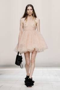 Model on a runway wearing a pale pink textured strapless mini dress with a voluminous ruffled hem, gold earrings, and black floral shoes.