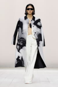 Model on a runway wearing a gray and black abstract coat over white wide-leg pants and a black top, with oversized sunglasses and statement earrings.
