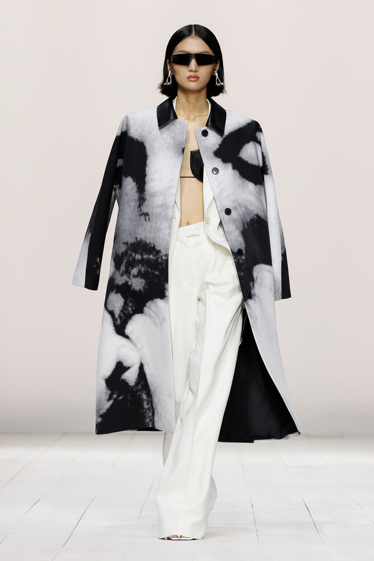 Model on a runway wearing a gray and black abstract coat over white wide-leg pants and a black top, with oversized sunglasses and statement earrings.