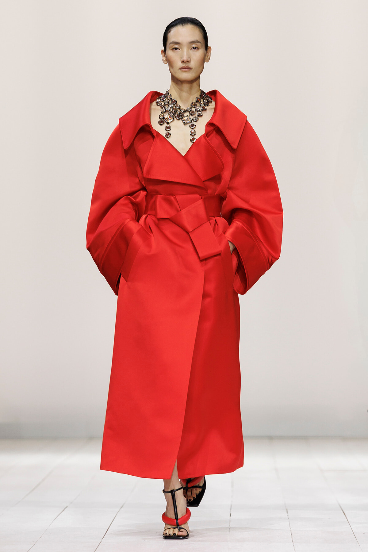Model on a runway wearing a bright red oversized coat with dramatic sleeves and a chunky gemstone necklace.]?
