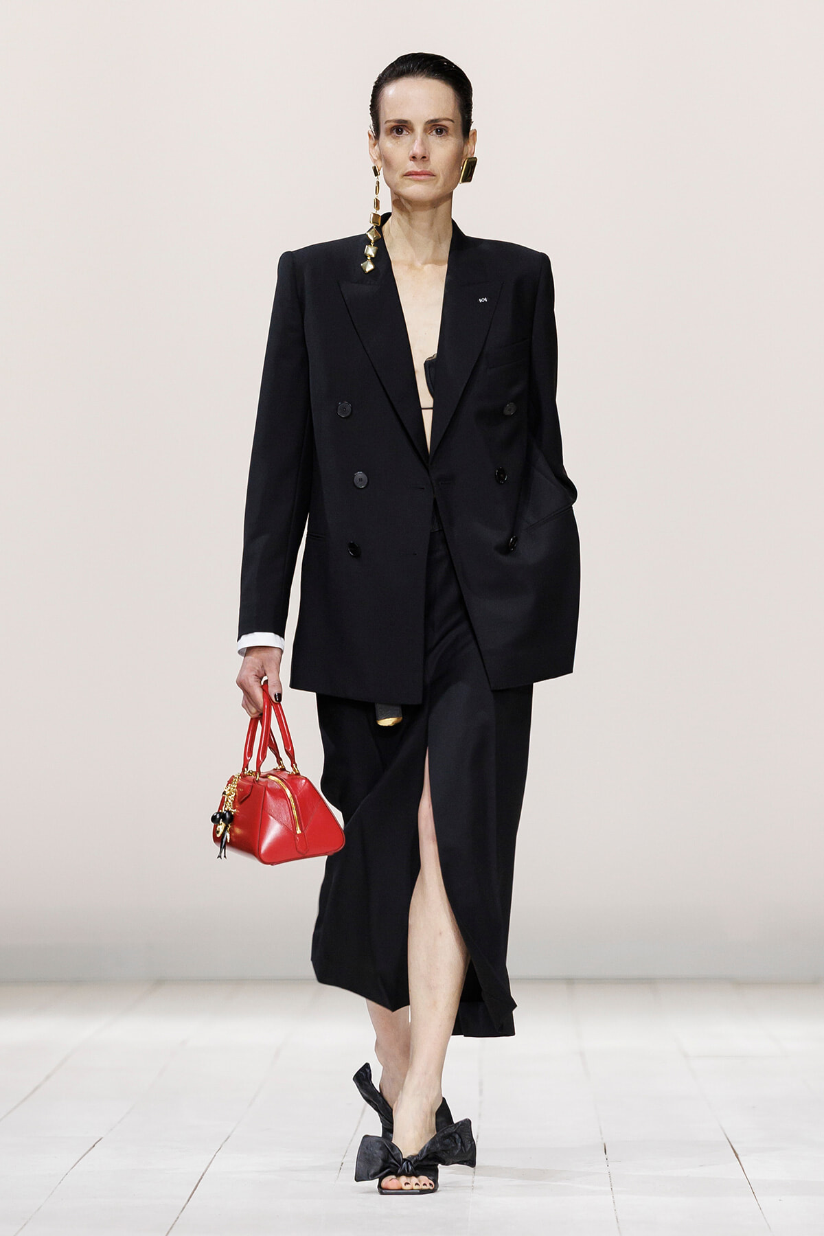Model on a fashion runway wearing a black double-breasted blazer dress with a front slit, carrying a red handbag.