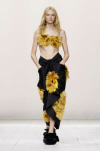 Model on the runway in a yellow feathered bandeau top and a black draped skirt with yellow feather appliques on the waist and hem.