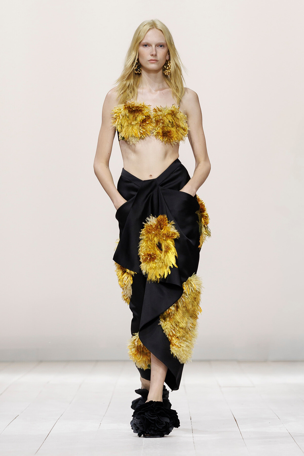 Model on the runway in a yellow feathered bandeau top and a black draped skirt with yellow feather appliques on the waist and hem.