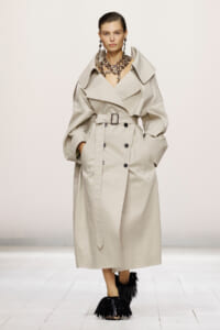 Model wearing a beige trench coat with a large collar and belt, accessorized with a chunky necklace on a runway.