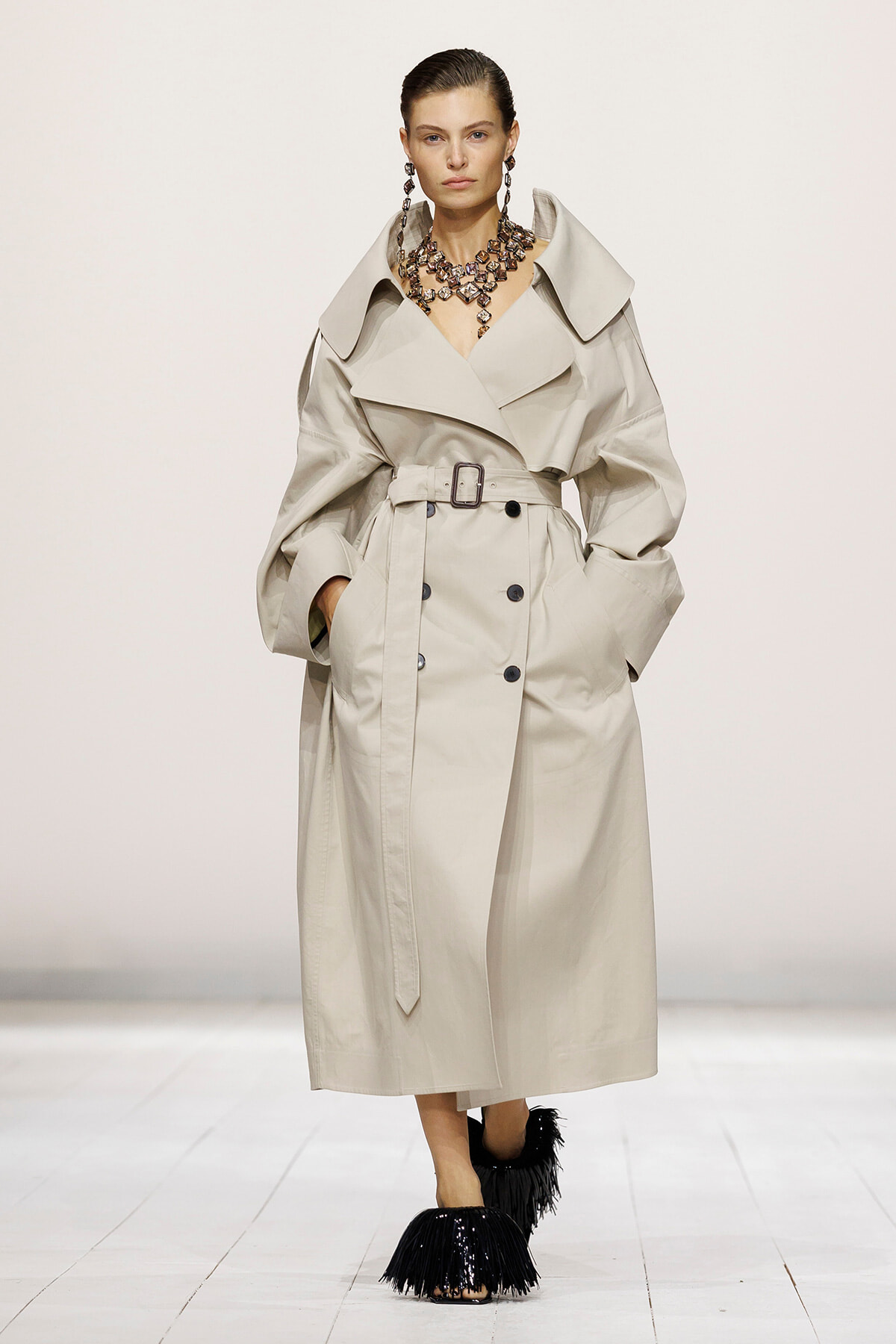 Model wearing a beige trench coat with a large collar and belt, accessorized with a chunky necklace on a runway.