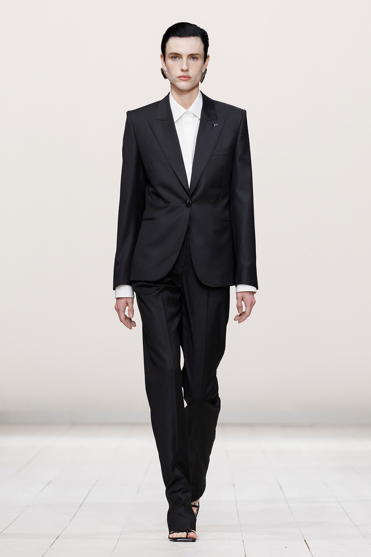 Female model in a black tailored pantsuit with a white shirt, walking down a runway in black heeled sandals.