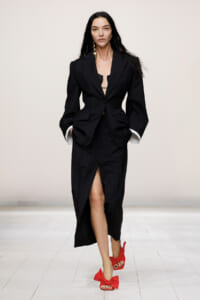 Runway model in a black tailored blazer with matching slit skirt, hands in pockets, red bow-embellished sandals.