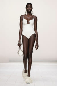 Model on a runway wearing a white textured one-piece swimsuit with a plunging neckline, oversized earrings, fringe sandals, and a small white handbag.