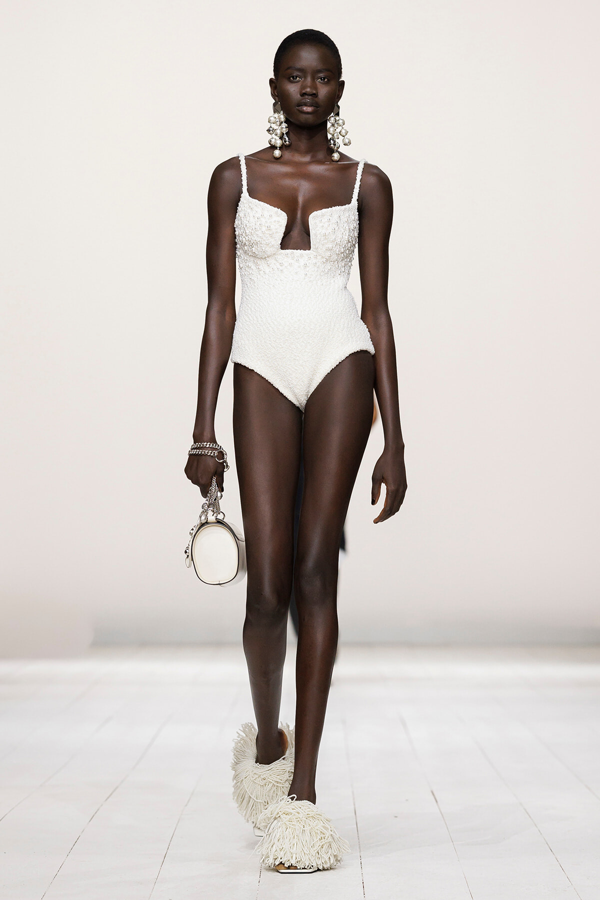 Model on a runway wearing a white textured one-piece swimsuit with a plunging neckline, oversized earrings, fringe sandals, and a small white handbag.