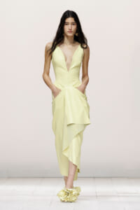 Model walking a runway in a pale yellow V-neck gown with a draped asymmetric skirt and pockets.