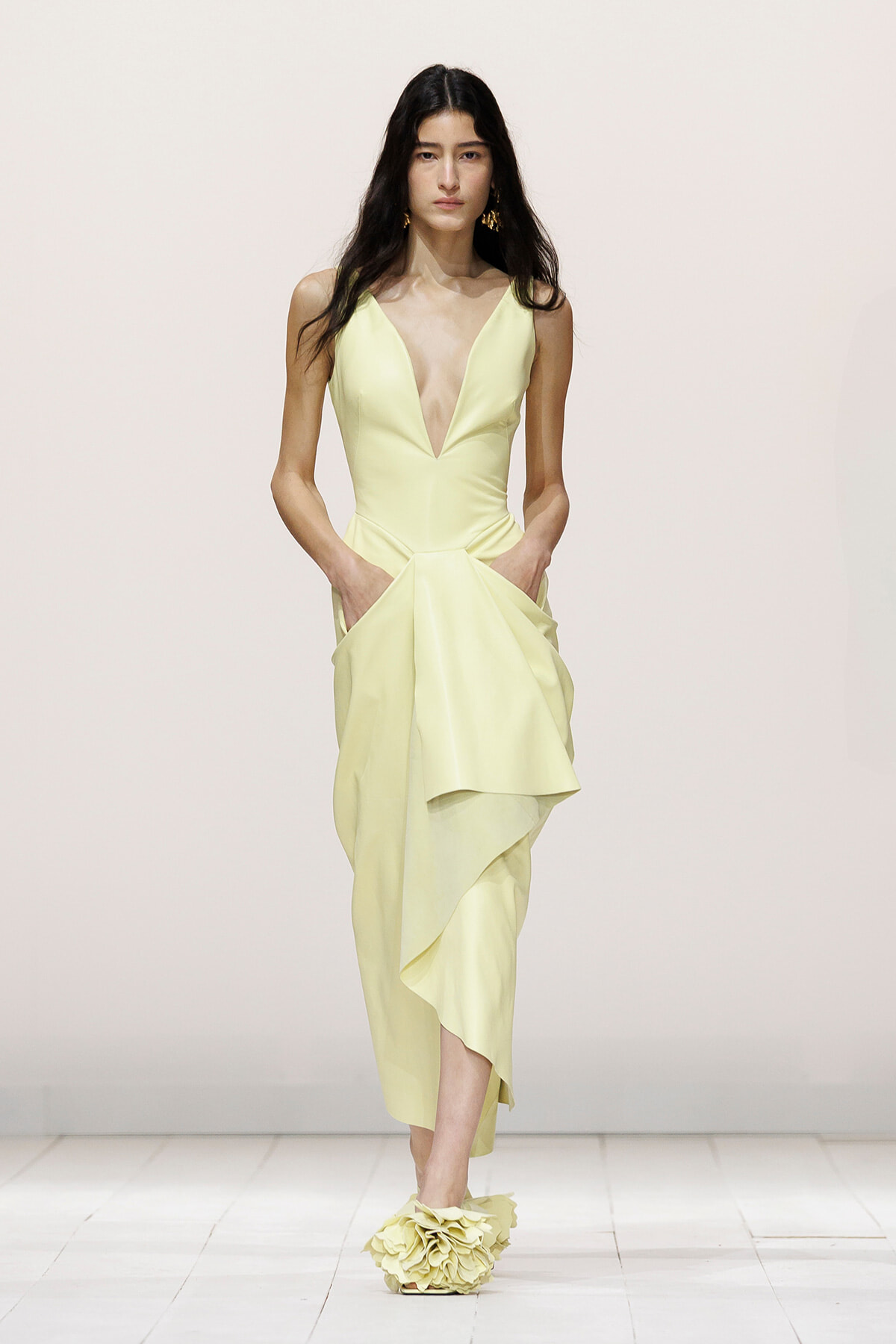 Model walking a runway in a pale yellow V-neck gown with a draped asymmetric skirt and pockets.