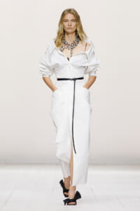 Model on a white runway wearing a white oversized shirt dress with a black belt, chunky crystal necklace, and black bow-strap heels.
