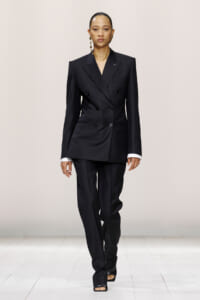 Model walking runway in a tailored black double-breasted blazer with matching wide-leg trousers and white cuffs showing, accessorized with bold earrings.