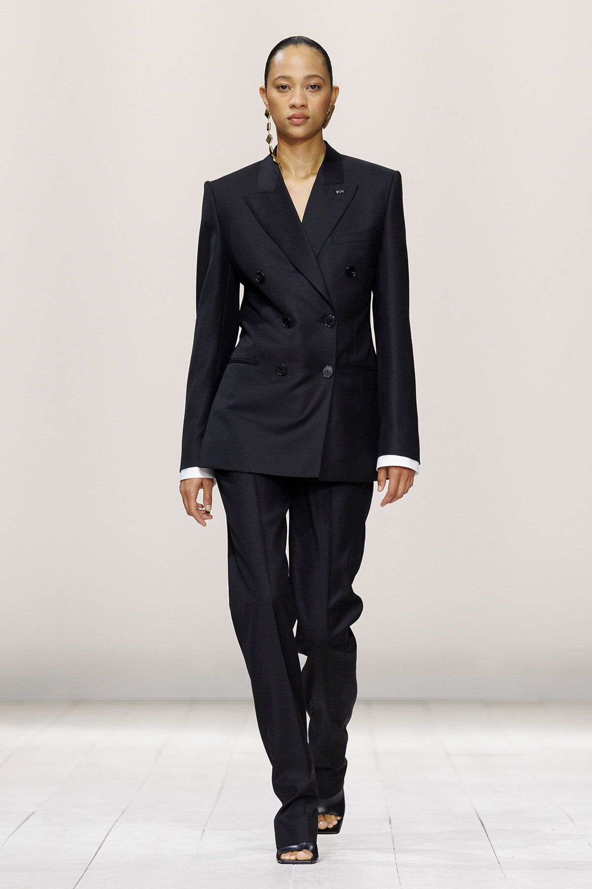 Model walking runway in a tailored black double-breasted blazer with matching wide-leg trousers and white cuffs showing, accessorized with bold earrings.