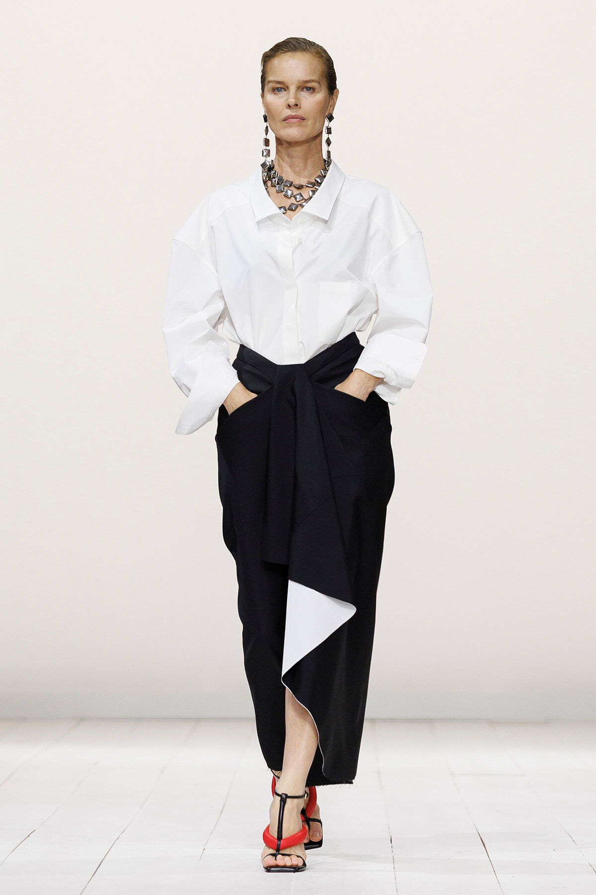 Model on a runway wearing an oversized white shirt and black draped wrap skirt with a front tie, paired with bold jewelry and red-strap sandals.