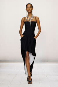 Model walking a runway in a black slip dress with an asymmetrical draped hem, layered pearl necklace, and black strappy heels.