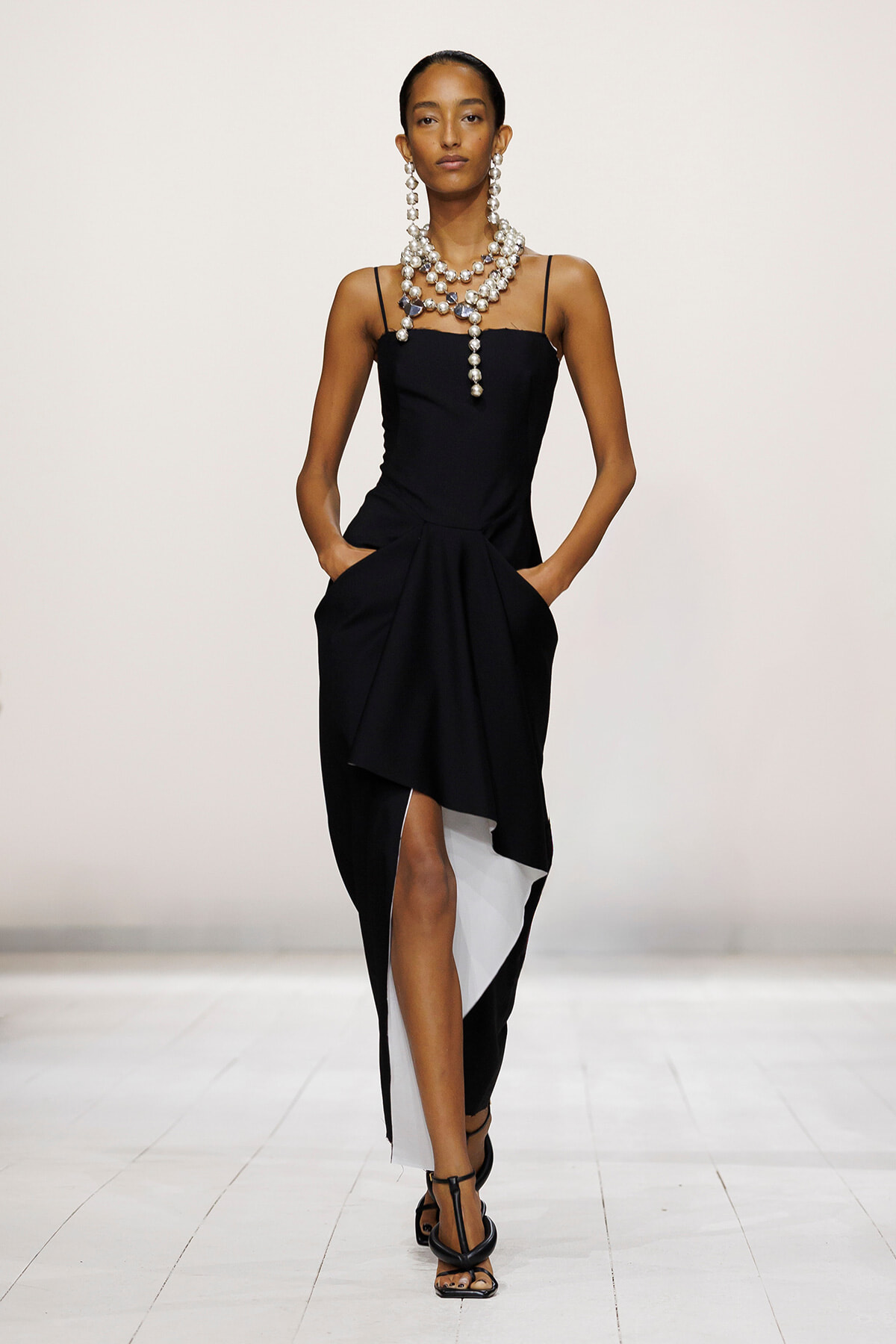 Model walking a runway in a black slip dress with an asymmetrical draped hem, layered pearl necklace, and black strappy heels.
