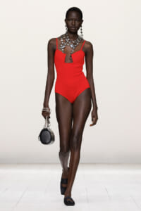 Runway model in a red one-piece swimsuit with a cutout neckline and chunky silver jewelry, carrying a small round black bag.