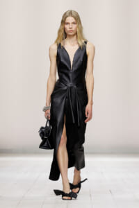 Runway model in a black satin, sleeveless gown with a plunging V-neck, sculpted waist, and high slit, carrying a small black handbag.
