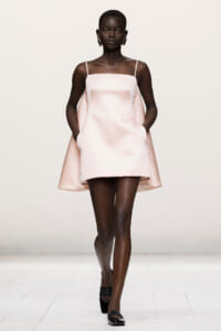 African woman model walks the runway in a pale pink mini dress with thin spaghetti straps and cape-like draped sides on a white studio backdrop.