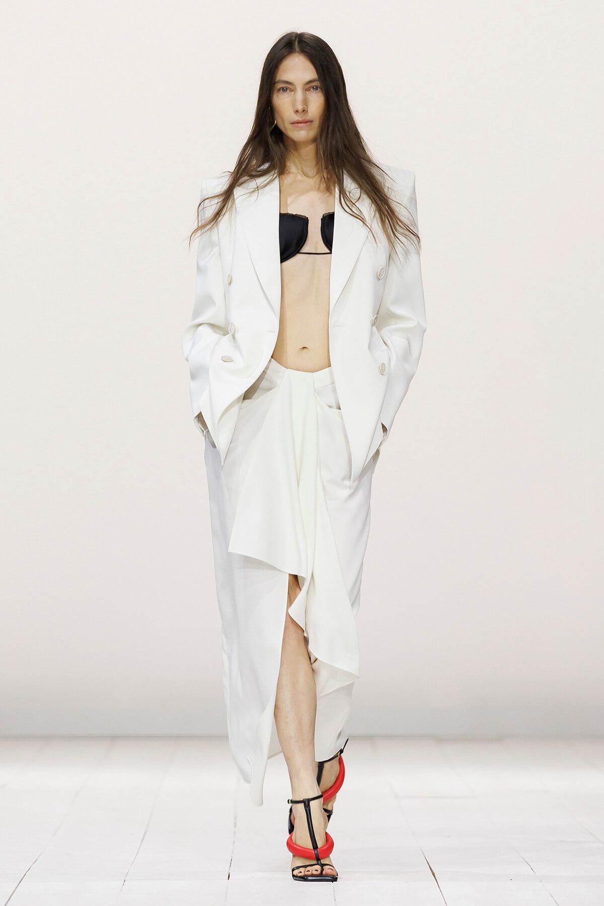 Model wearing an oversized white suit with a black bralette, hands in pockets, white skirt with a front slit, red strappy heels on a runway.