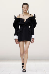 Runway model in a black off-shoulder mini dress with dramatic oversized sleeves, white cuffs, long geometric earrings, and black heels.