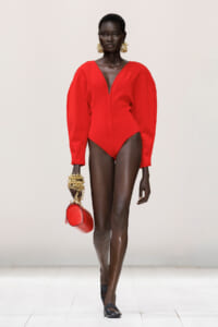 Model walking a runway in a bold red long-sleeve bodysuit with a deep V at the neckline, paired with gold statement earrings and matching red purse.