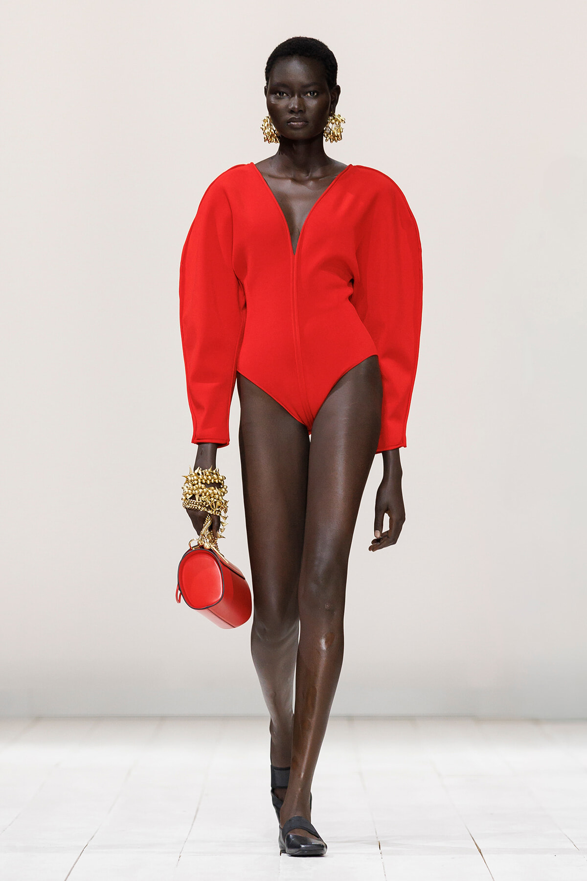 Model walking a runway in a bold red long-sleeve bodysuit with a deep V at the neckline, paired with gold statement earrings and matching red purse.