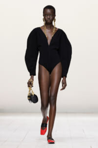 Model walking a fashion runway in a black deep-V bodysuit with oversized sleeves, gold jewelry, and a small round black purse on a gold chain, red-black heels.