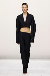 Model on a white runway wearing a black cropped blazer and matching high-waisted trousers, midriff exposed on a fashion show runway.