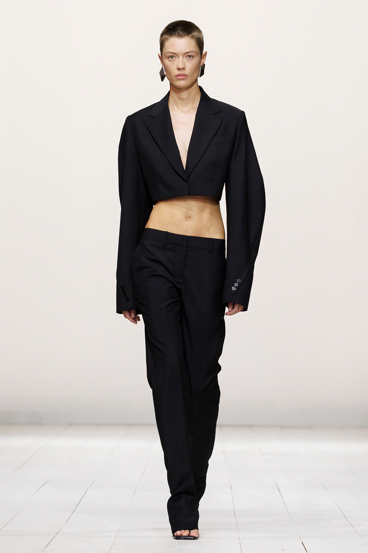Model on a white runway wearing a black cropped blazer and matching high-waisted trousers, midriff exposed on a fashion show runway.