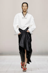 Model on a runway wearing an oversized white shirt paired with a black asymmetric draped leather skirt and red-black strappy sandals, with bold jewelry.