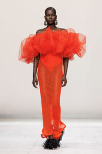 Model on a runway wearing a striking bright orange off-shoulder gown with a large voluminous mesh ruffle collar and a dotted mesh skirt with feathered black shoes.