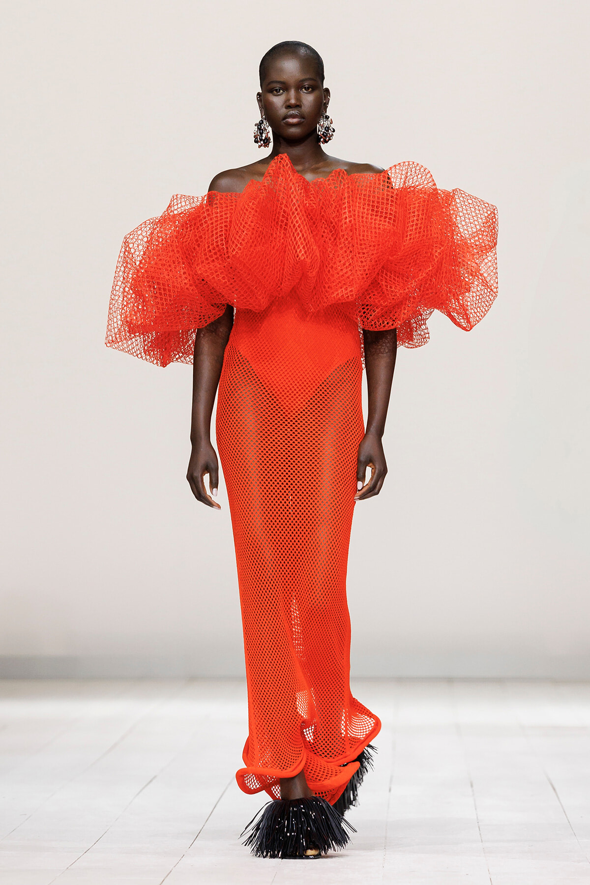 Model on a runway wearing a striking bright orange off-shoulder gown with a large voluminous mesh ruffle collar and a dotted mesh skirt with feathered black shoes.