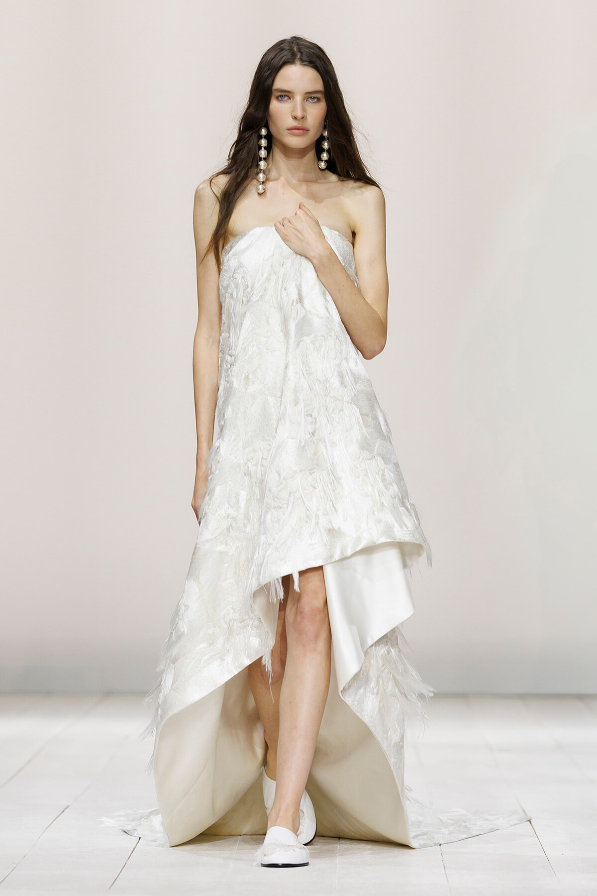 Model walking the runway in a strapless white textured high-low gown with oversized pearl earrings.