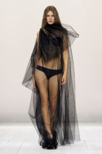 Model walking a fashion show in a long black mesh cape over a black bikini on a white runway.