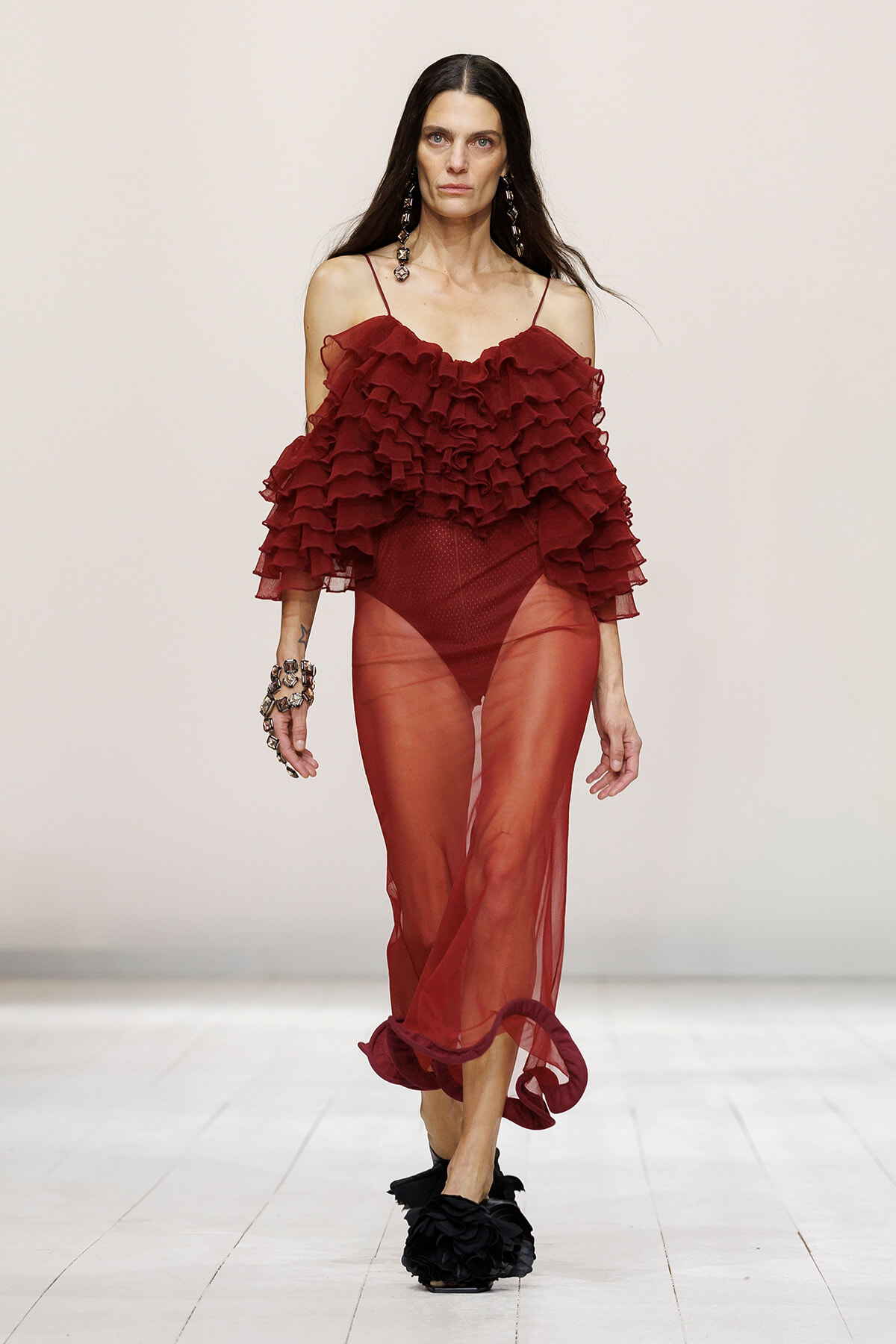 Model walks the runway in a burgundy, off-the-shoulder, multi-layered ruffled top with a sheer burgundy skirt and black floral-embellished shoes.