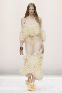 Model walks the runway in a cream off-the-shoulder ruffled top, sheer pants, and large cream floral sandals.