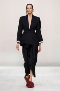 Model on a runway wearing a black tailored blazer with a plunging V, a layered peplum skirt, white cuffs, bold square earrings, and burgundy fringe shoes.