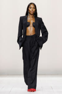 Model walks the runway in an oversized black suit with a cropped bralette, long dangling earrings, and red fringe shoes.