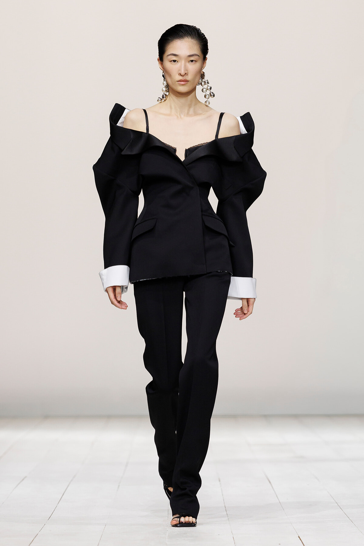 Runway look: a female model in a black off-shoulder structured blazer with dramatic shoulders, white cuffed sleeves, and matching black pants.