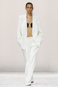 Model walks the runway in a white oversized suit, blazer open to reveal a black bra and long statement earrings.