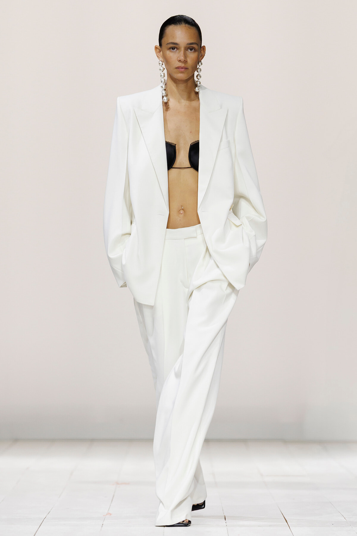 Model walks the runway in a white oversized suit, blazer open to reveal a black bra and long statement earrings.