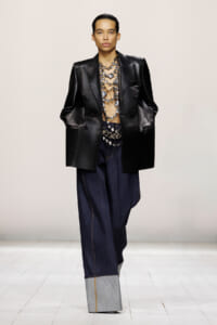 Model on a runway wearing an oversized black leather blazer, sheer top with metallic necklace, and wide-leg dark denim jeans with a light gray cuff.