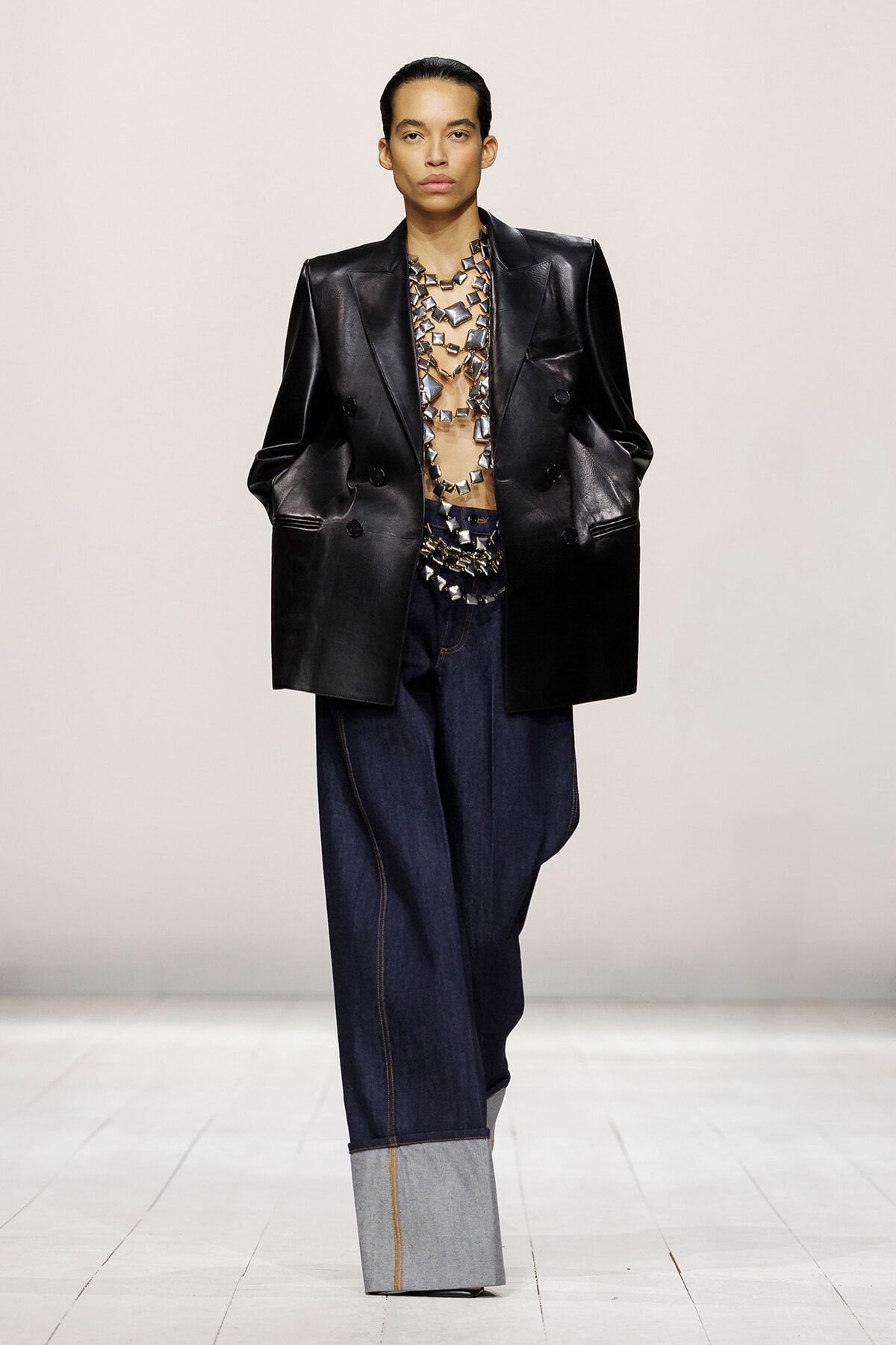 Model on a runway wearing an oversized black leather blazer, sheer top with metallic necklace, and wide-leg dark denim jeans with a light gray cuff.