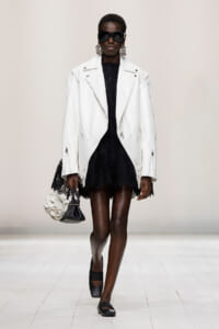 Model on a runway wearing an oversized white leather jacket over a black lace dress, with large sunglasses, statement earrings, and a small black-and-white handbag.