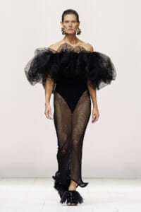 Runway model wearing an off‑the‑shoulder black ruffle top with voluminous mesh sleeves and black fishnet tights.