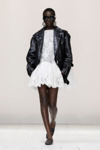 Model on runway wearing a black oversized leather jacket over a white lace mini dress, with large sunglasses and statement earrings.