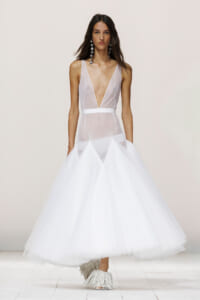 Model wearing a white mesh-plunge gown with a deep V bodice, belt, and voluminous tulle skirt on a runway.